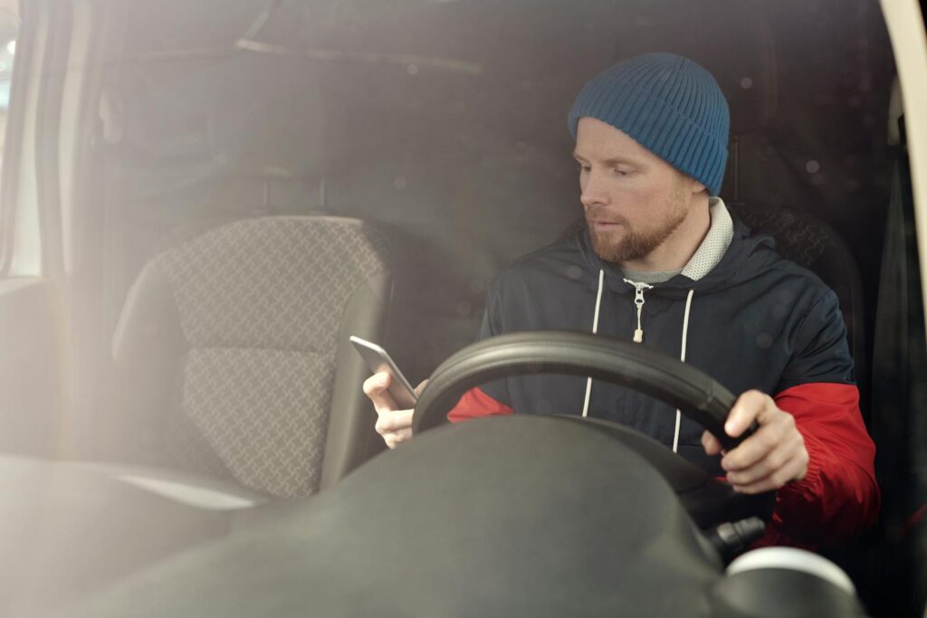 A man in a beanie uses his smartphone while sitting in a car. Daylight setting.