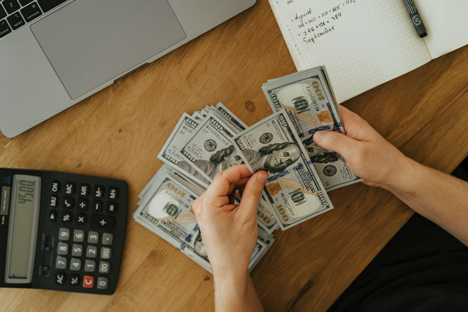 Close-up of hands counting dollar bills with calculator, notebook, and laptop on wooden table.