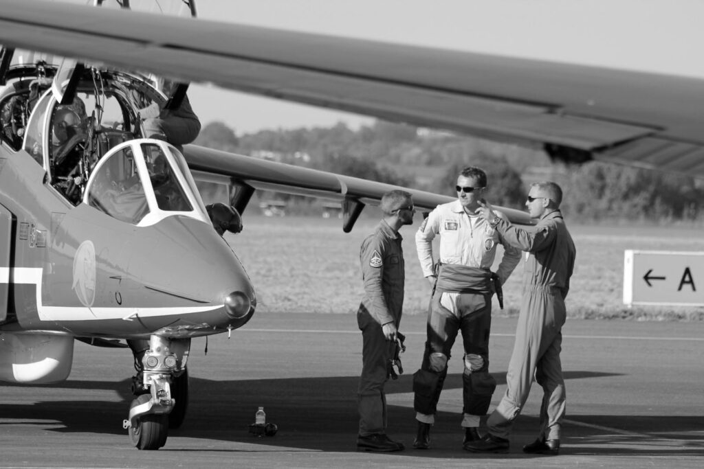 Three military pilots in conversation beside a jet on a sunny day at an airfield.