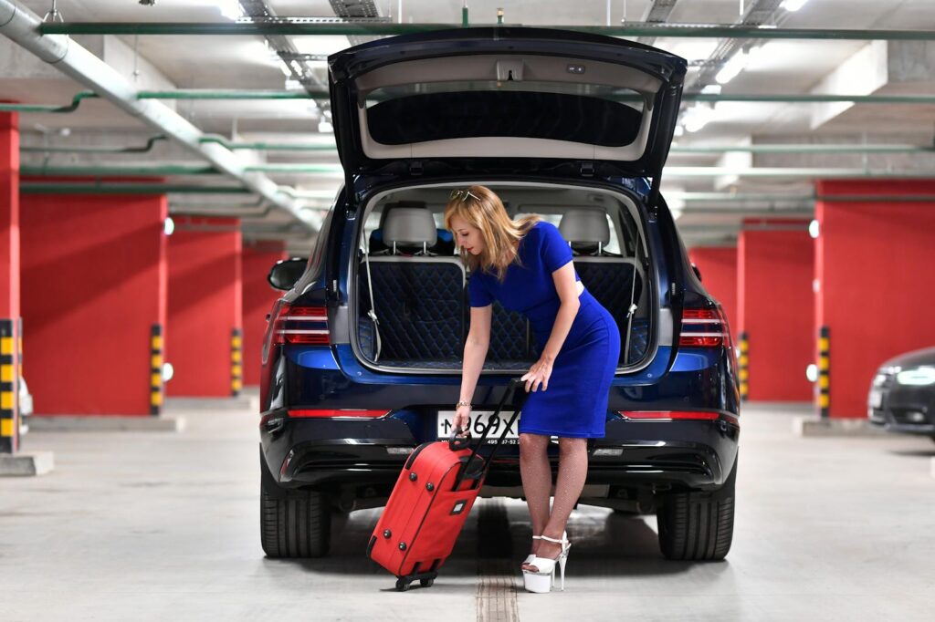 Stylish woman in a blue dress placing luggage in car trunk in a parking garage.