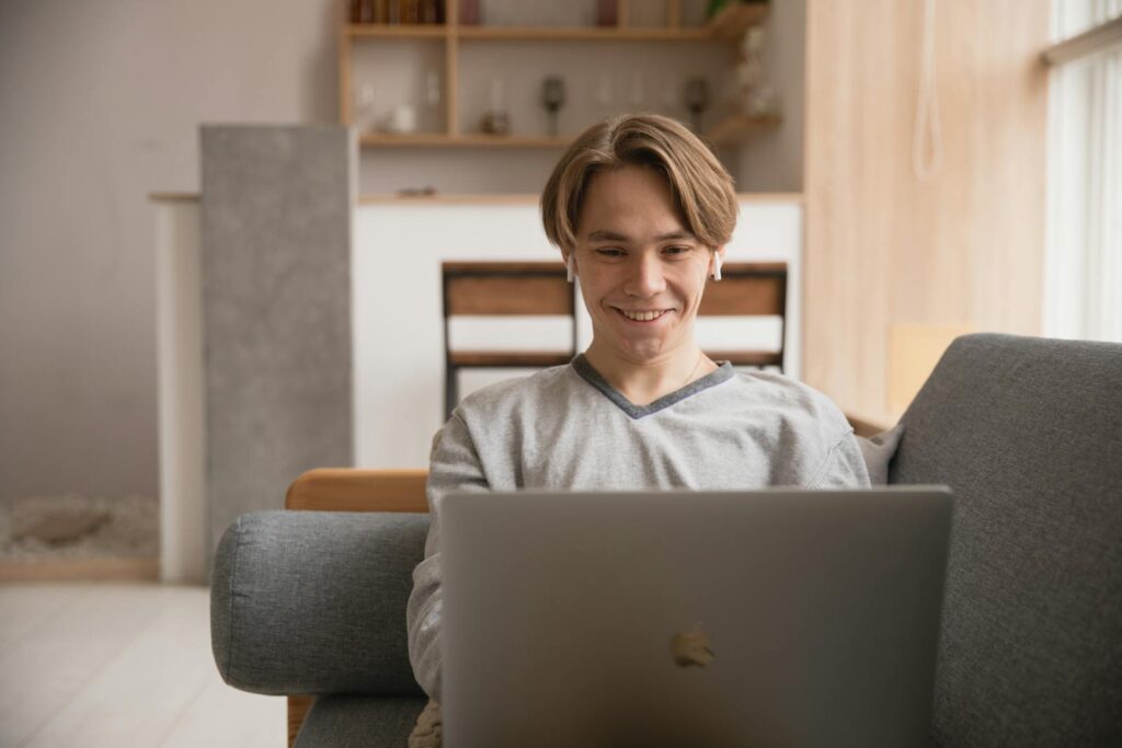 Smiling young man using a laptop on a couch at home, symbolizing remote work and digital lifestyle.