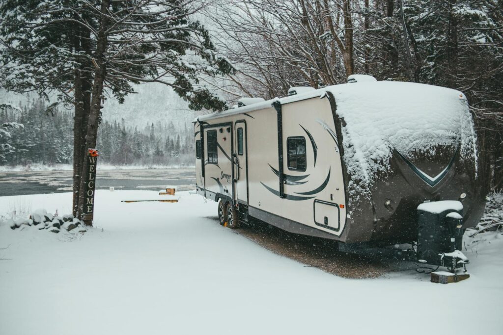 Travel trailer covered with snow placed on shore near river in winter forest in countryside in daylight