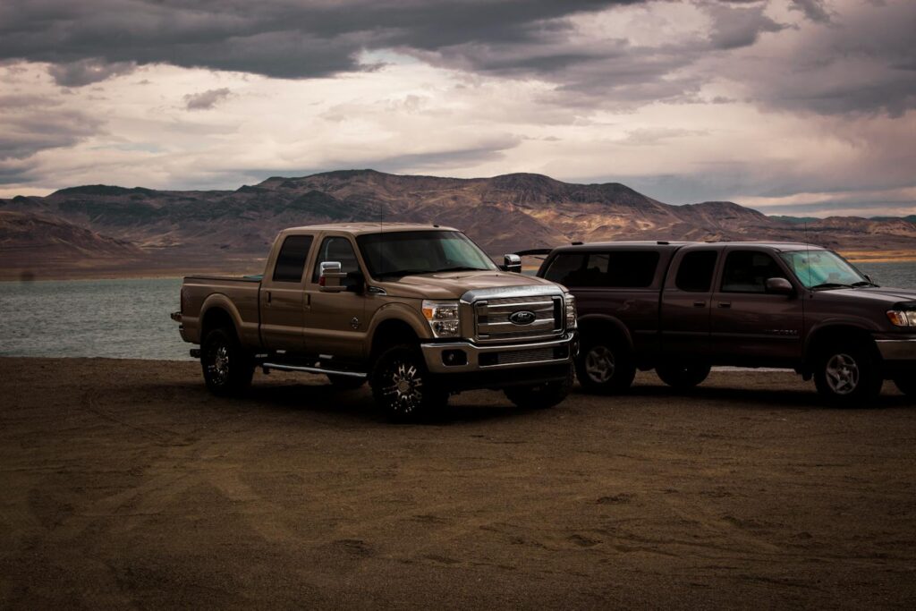 Pickup trucks parked by a serene mountain lake under a cloudy sky, creating a rugged, adventurous scene.