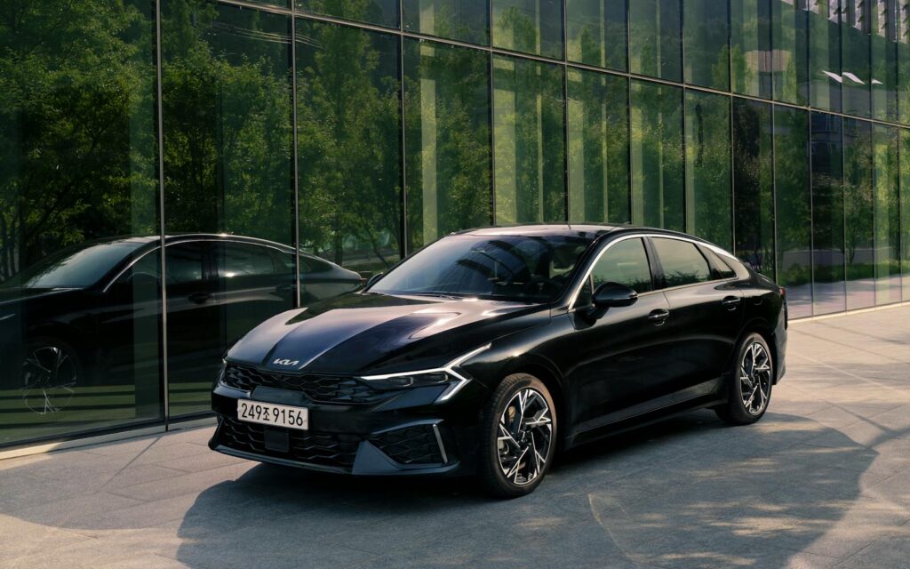 A stylish black Kia K5 sedan parked in a modern urban area, reflecting city architecture.