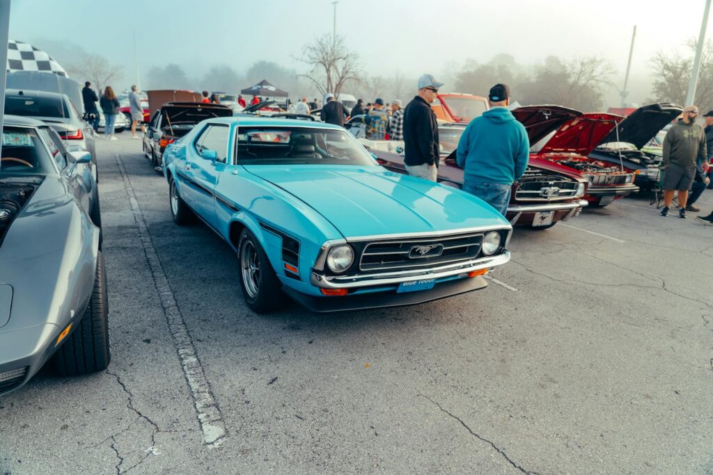 Classic blue Mustang displayed at an outdoor car show with enthusiasts gathered around.