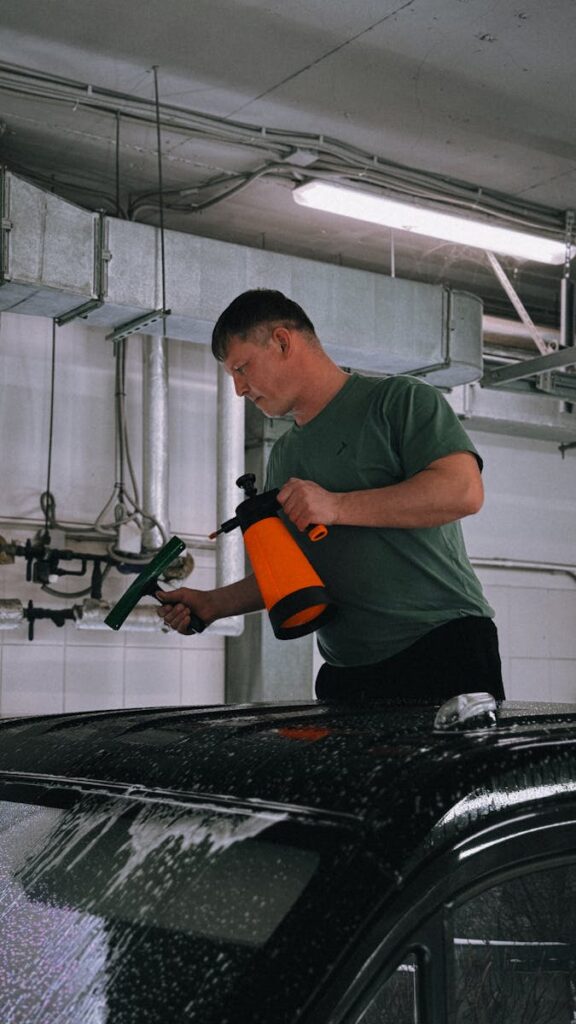 A man cleans a car's roof with a spray bottle and tool in an indoor garage.