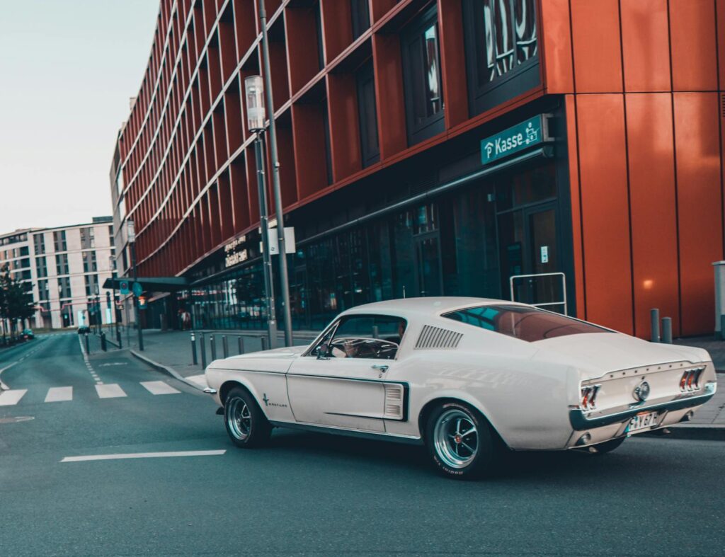 Classic white Mustang cruising past modern urban architecture on a city street.