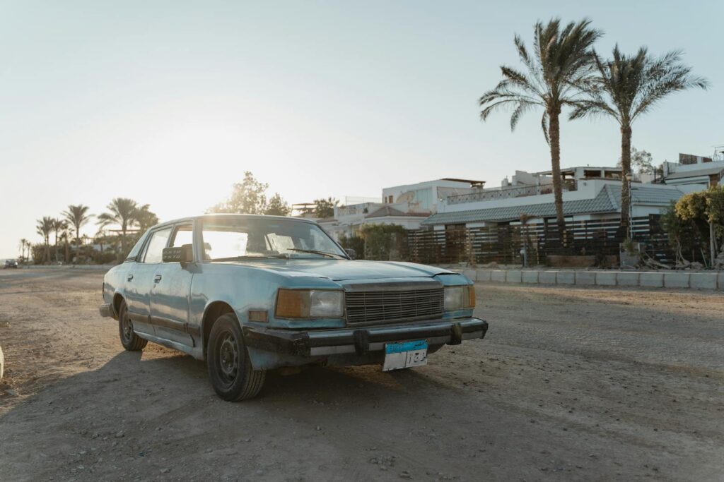 Classic car with aging details parked on a rural unpaved road with palm trees at sunset.