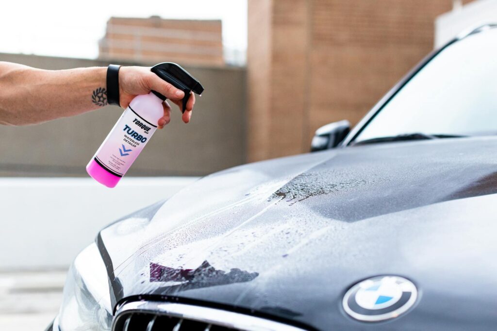 Hand applying cleaning spray on a wet BMW hood for car detailing.