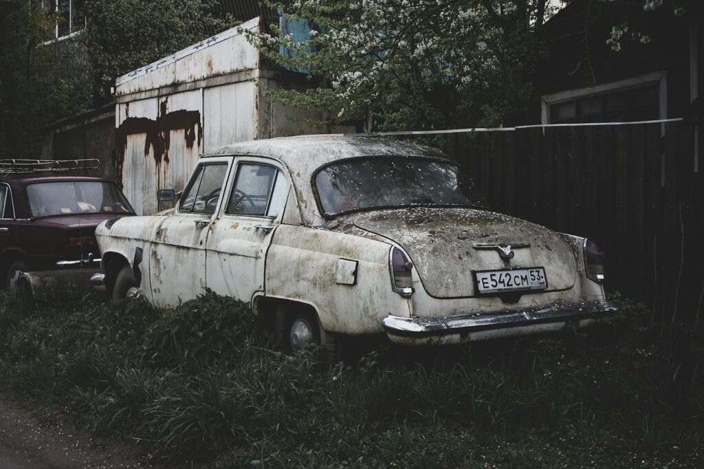 A weathered vintage car sits abandoned in an overgrown yard in Veliky Novgorod, Russia.