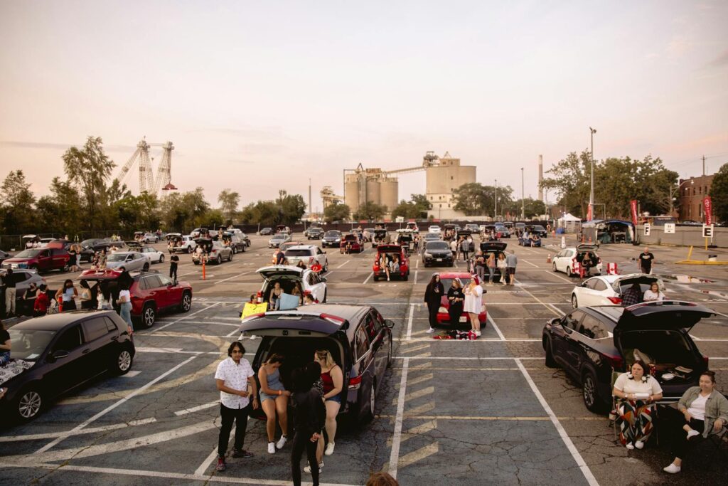 People gathering around cars in a spacious parking lot under a clear sky.