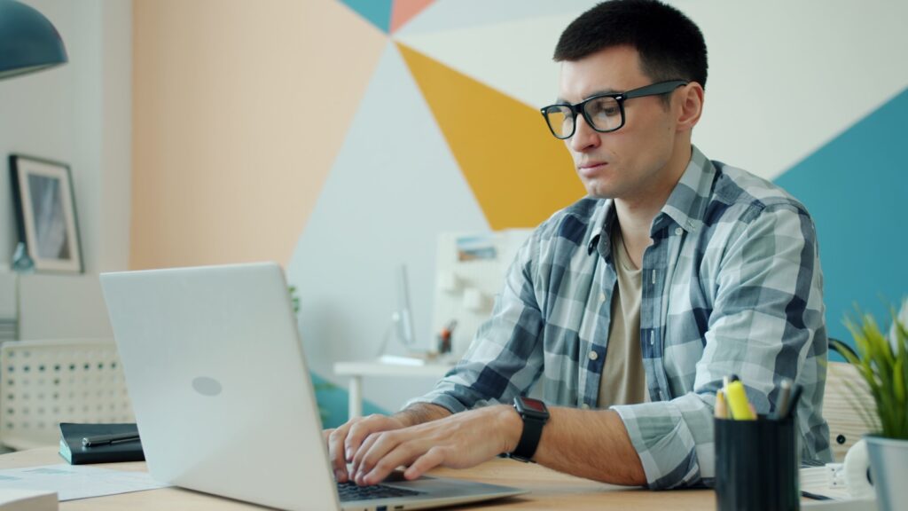 Man wearing glasses types on a laptop at a desk.