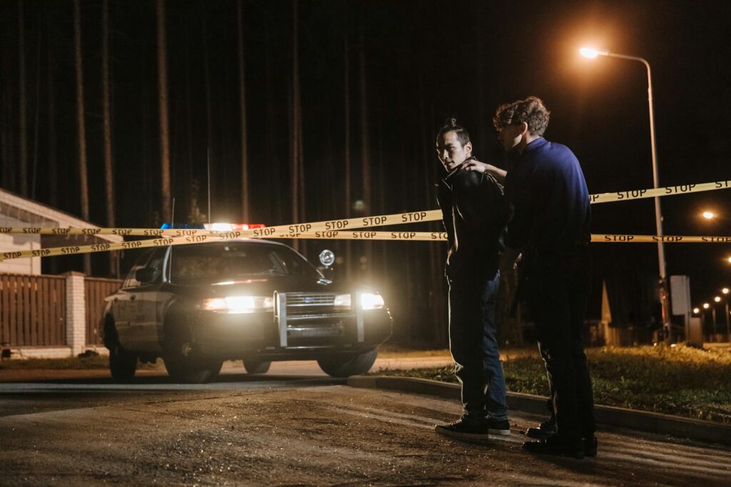 Police officer detaining a suspect near a crime scene at night with tape and patrol car.