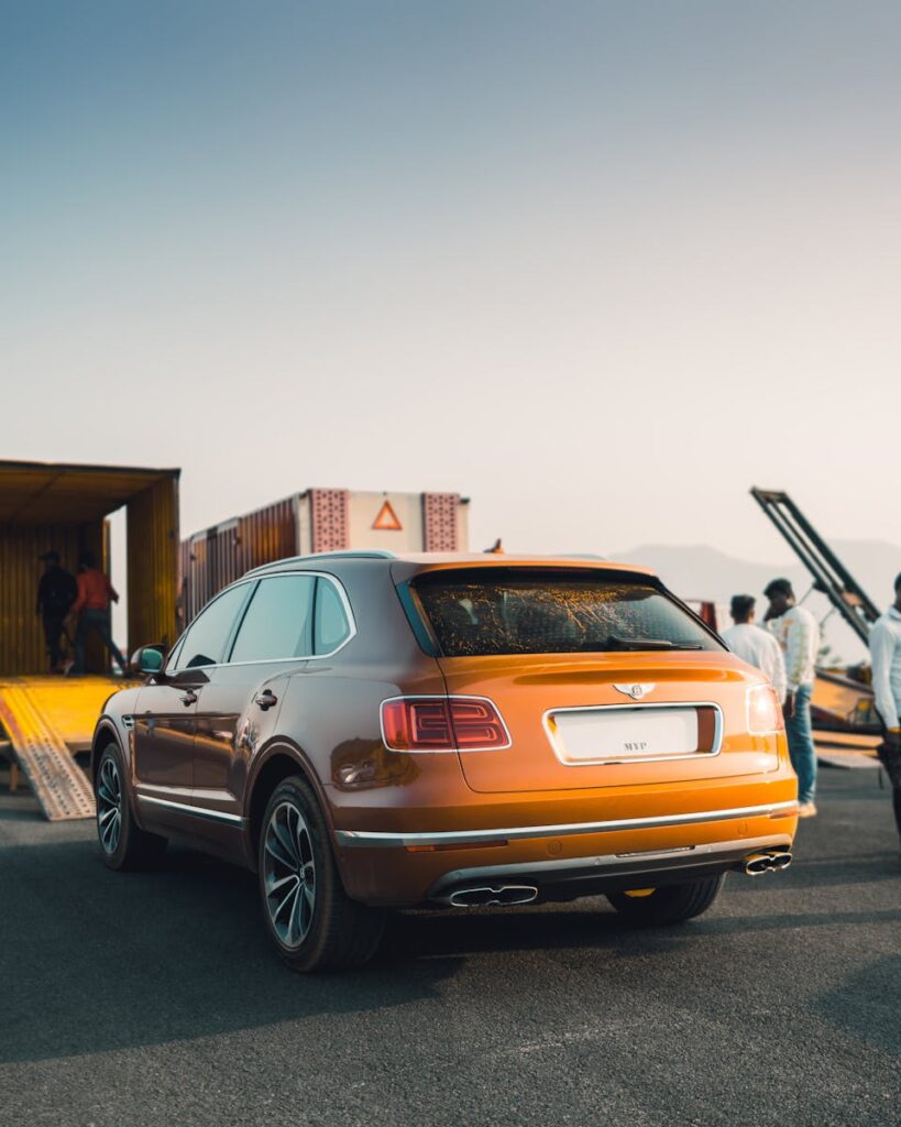 A sleek orange Bentley SUV near shipping containers under a clear sky, capturing elegance and adventure.