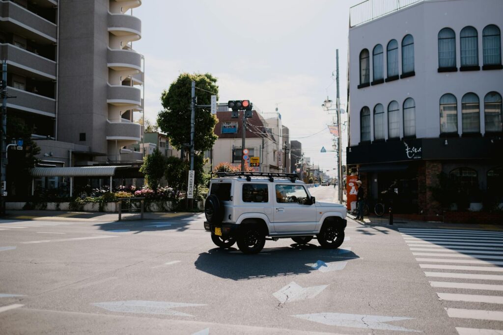 A Suzuki Jimny at a Tokyo intersection under sunlight, showcasing city life and urban architecture.