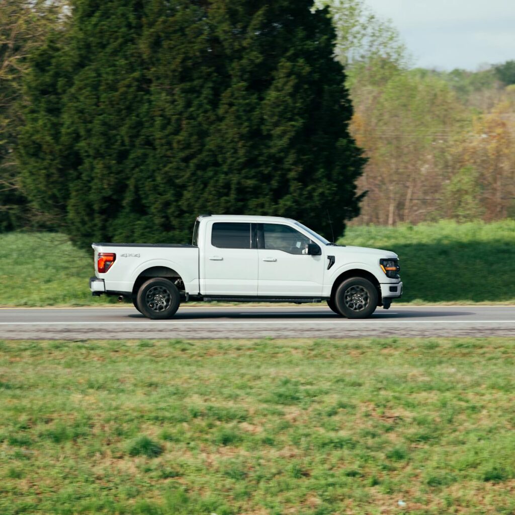 White pickup truck driving along a countryside road with lush greenery in the background.