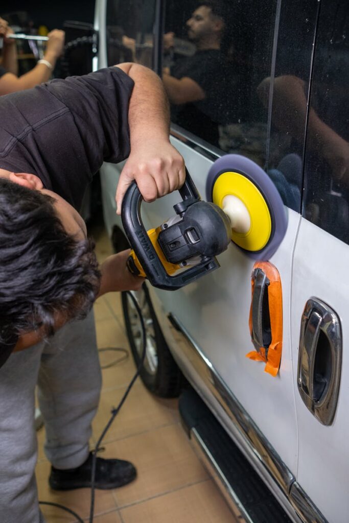 A man uses an electric buffer to polish and clean a white car door in a garage setting.