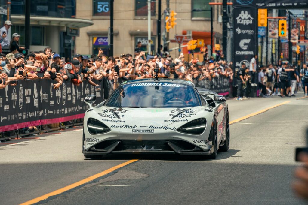 Supercar driving on city street during the Gumball 3000 race, surrounded by enthusiastic crowd.