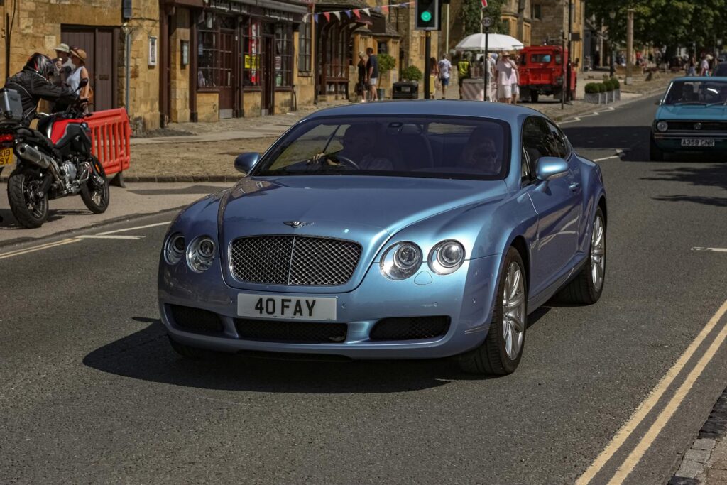 Blue Bentley Continental driving on a sunny city street with pedestrians and motorcycles.
