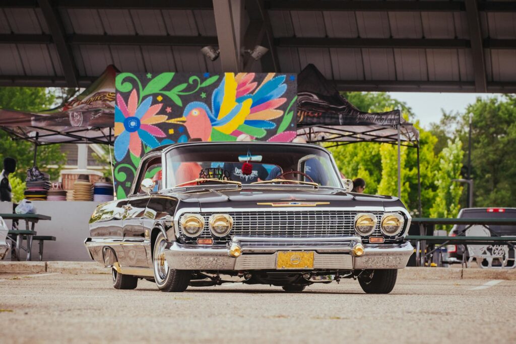 Vintage car parked at an outdoor market with vibrant mural backdrop, creating a lively urban scene.
