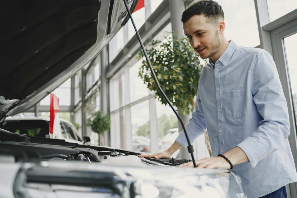 Young man examines car engine in a modern showroom, wearing a blue shirt.