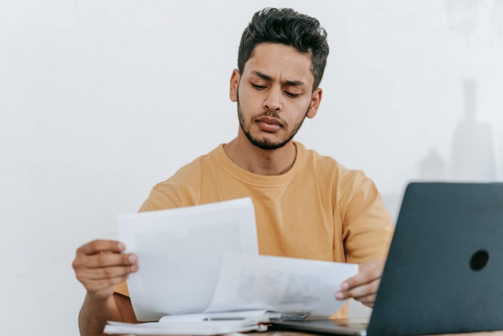 Attentive young bearded Hispanic male remote worker examining papers with results of project while working with laptop in home office