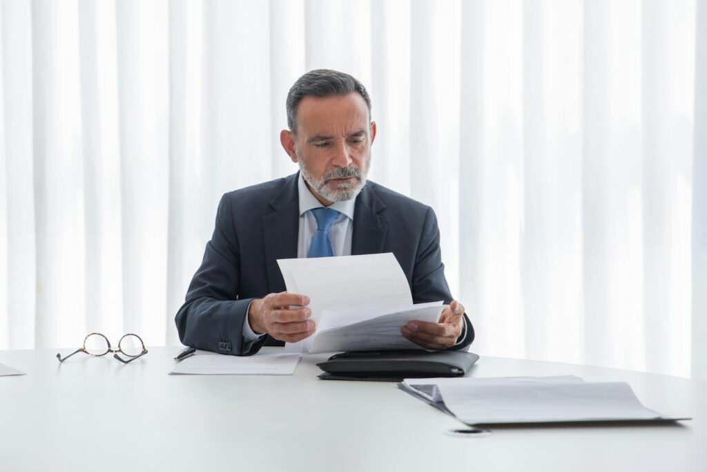 Elderly businessman focusing on reading documents at a table in a bright office setting.
