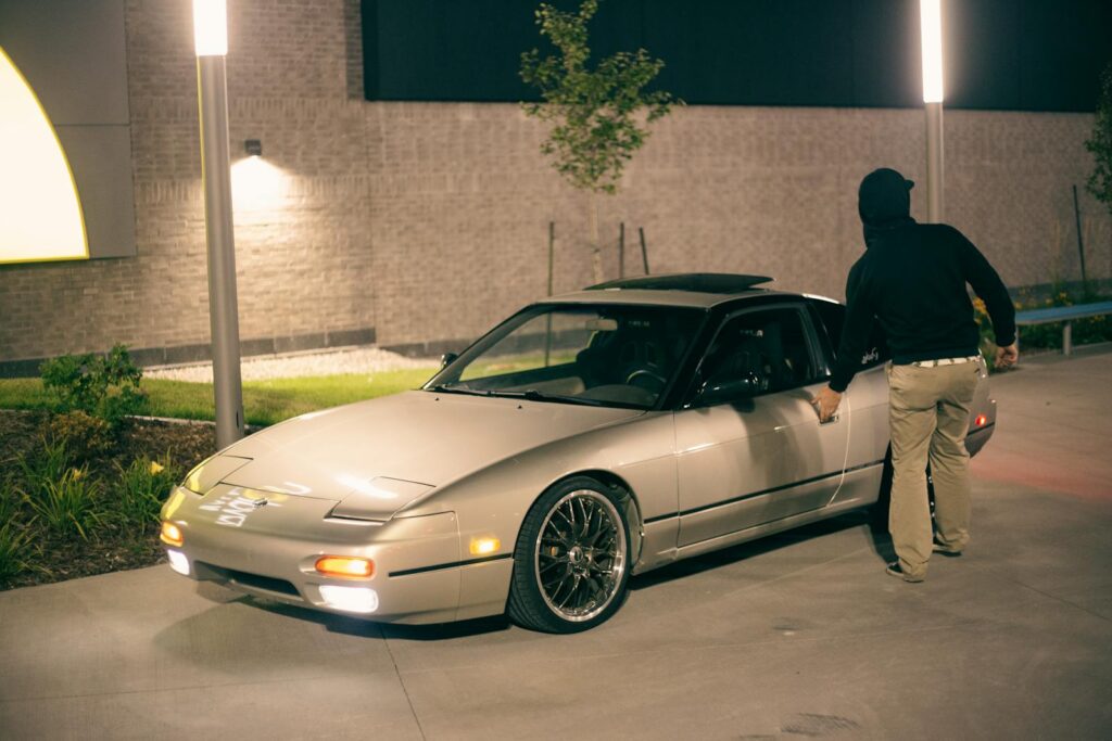 A man in a hoodie opens a car door under street lights at night, creating a mysterious urban vibe.