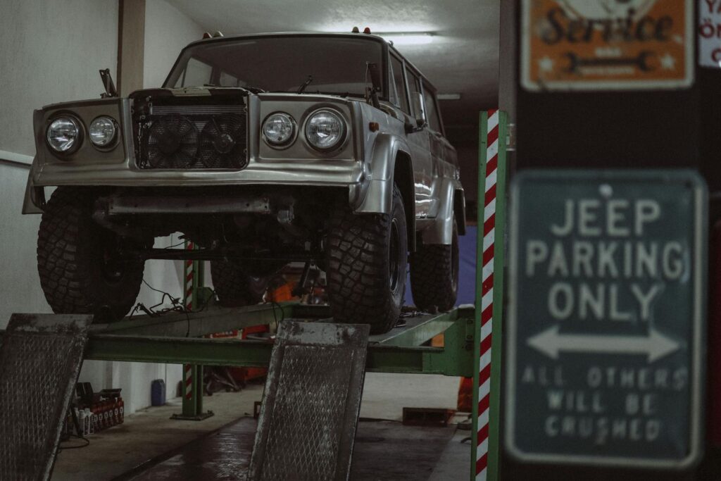 Classic Jeep on a lift in an indoor auto repair shop with a humorous parking sign.