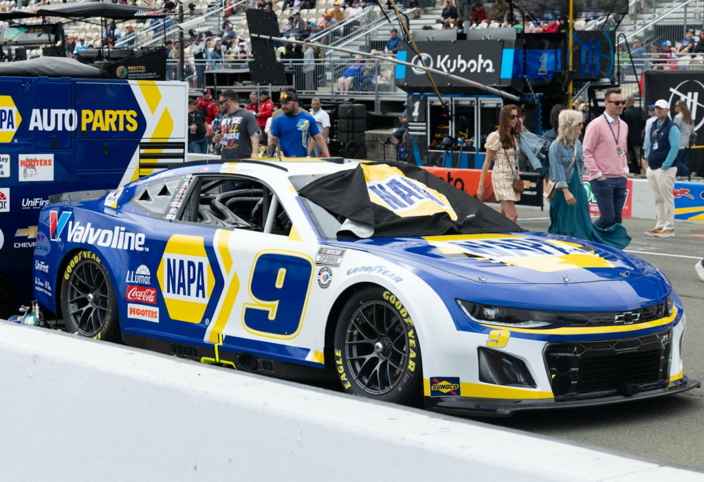 NASCAR race car in NAPA blue livery with branding, parked in a busy paddock area.
