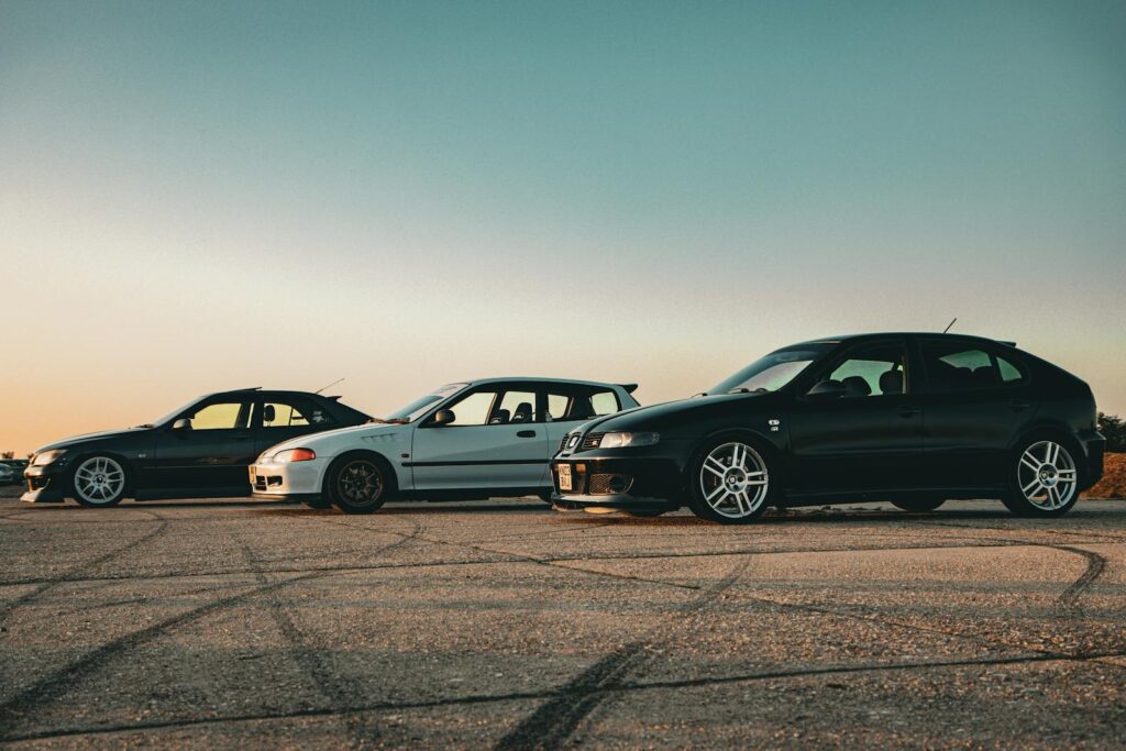 Three tuned sports cars parked on asphalt at sunset, showcasing vehicle customization and style.