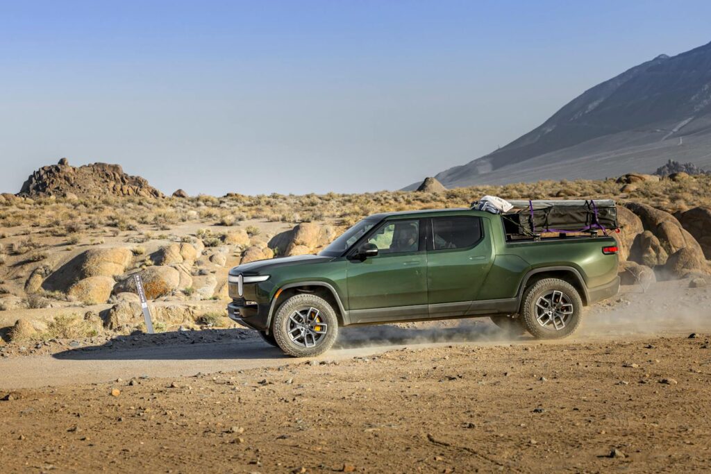 Green electric pickup truck on a dusty trail in a rocky desert landscape.
