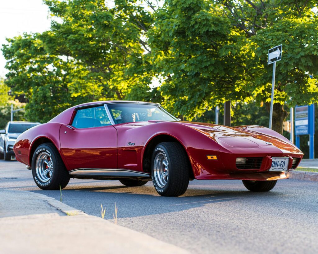 A vintage red sports car shines in the sunlight on a leafy street, showcasing timeless elegance.