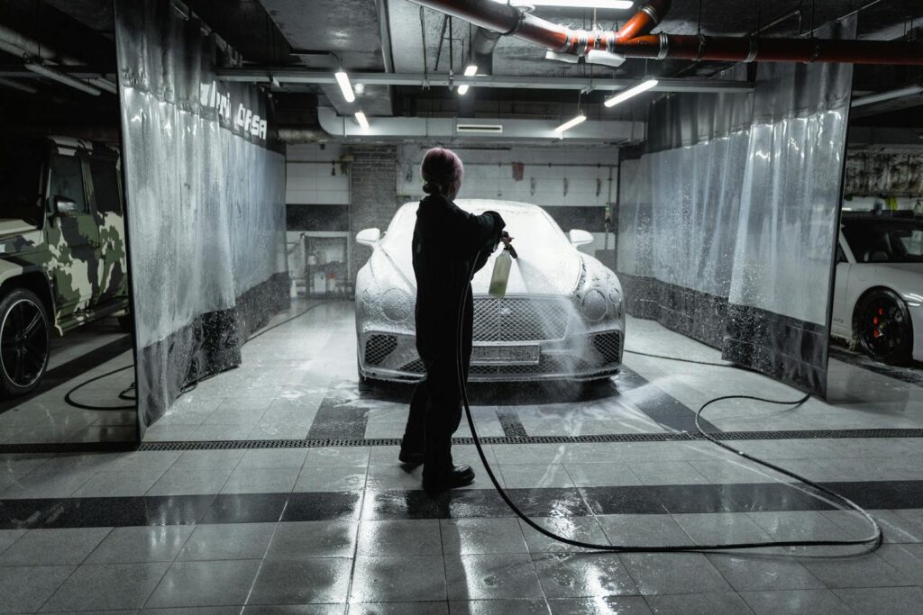 A person using a hose to spray a car with snow foam in an indoor carwash setting.