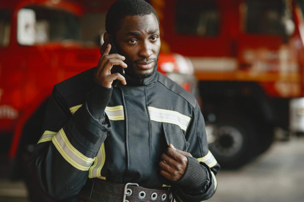 Close-up of a firefighter in protective gear making a phone call by a fire truck.