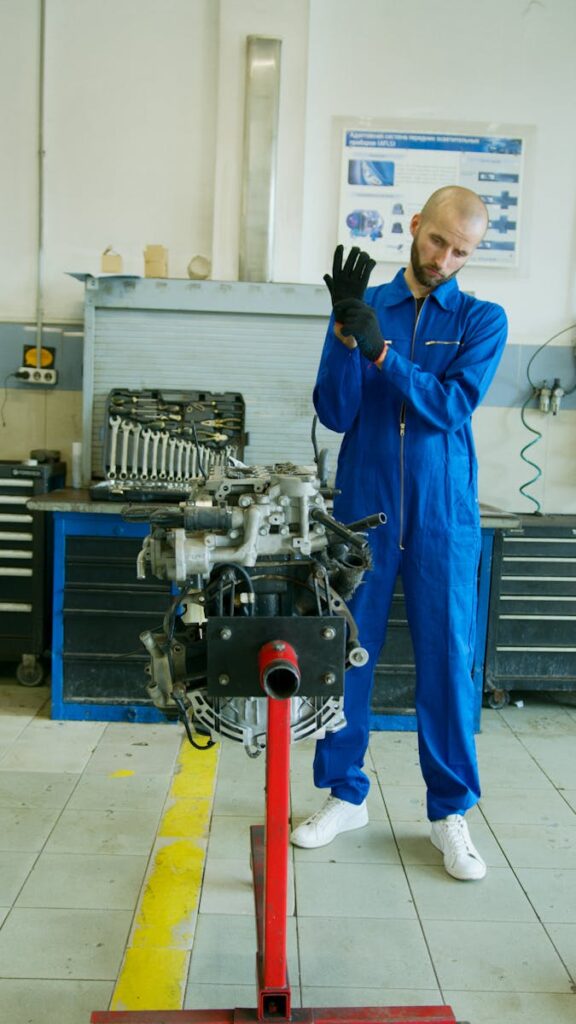 Mechanic in blue overalls adjusting gloves by engine in garage setting.