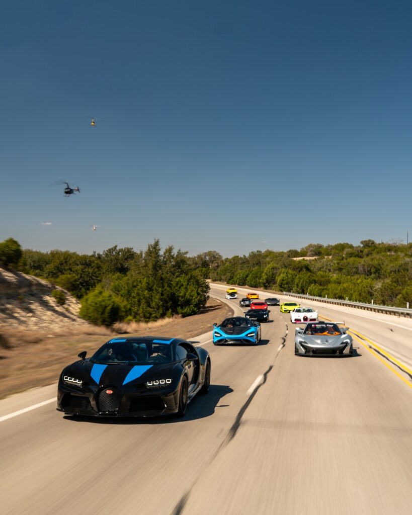 Luxury cars cruise down a sunny road.