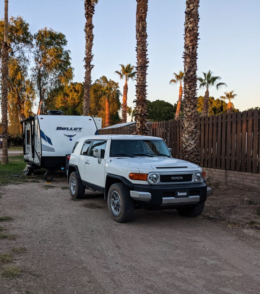 A white Toyota FJ Cruiser parked next to an RV under tall palm trees in Winterhaven, California.