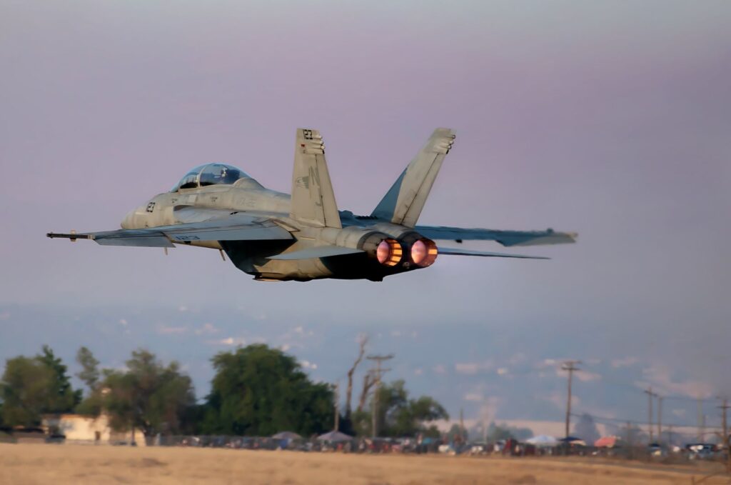 A military jet with afterburners activated takes off from a runway against the backdrop of a clear sky.