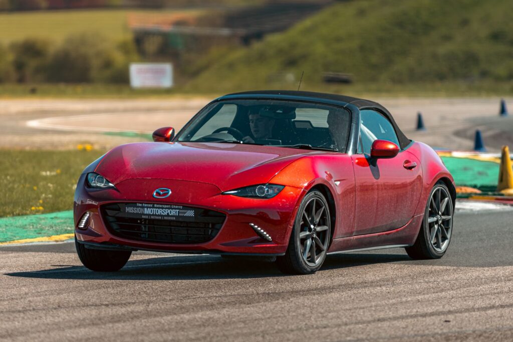 Red Mazda MX-5 sports car cornering on a track in England, under clear skies.