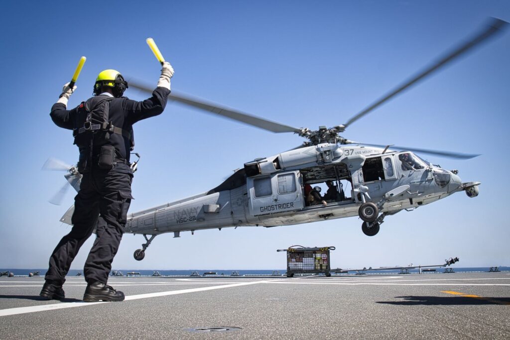 A military helicopter lands on an aircraft carrier guided by a deck crew member.