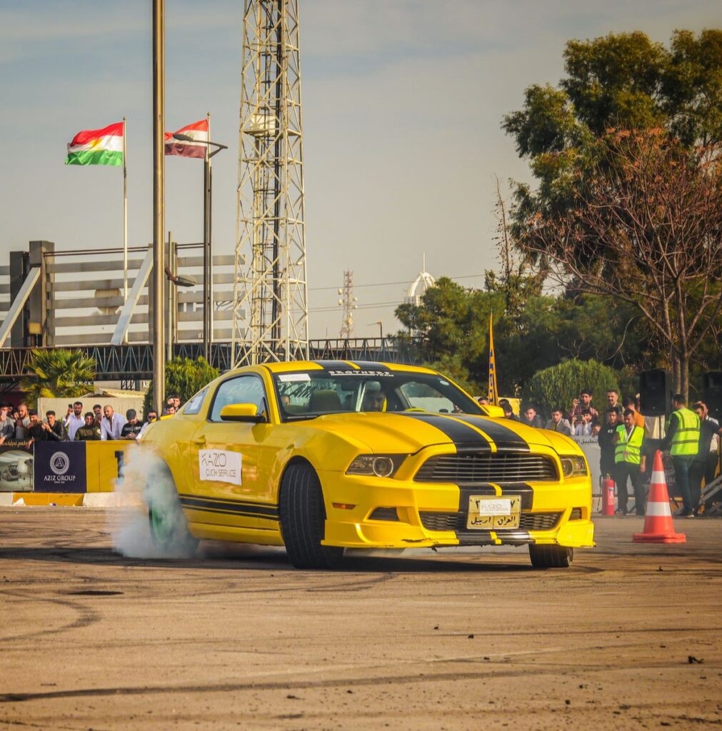 A dynamic shot of a yellow Ford Mustang drifting at an outdoor car show with flags waving.