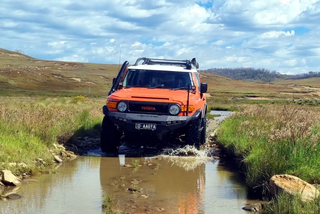 An orange SUV driving through rocky terrain with water and grass under a cloudy sky.