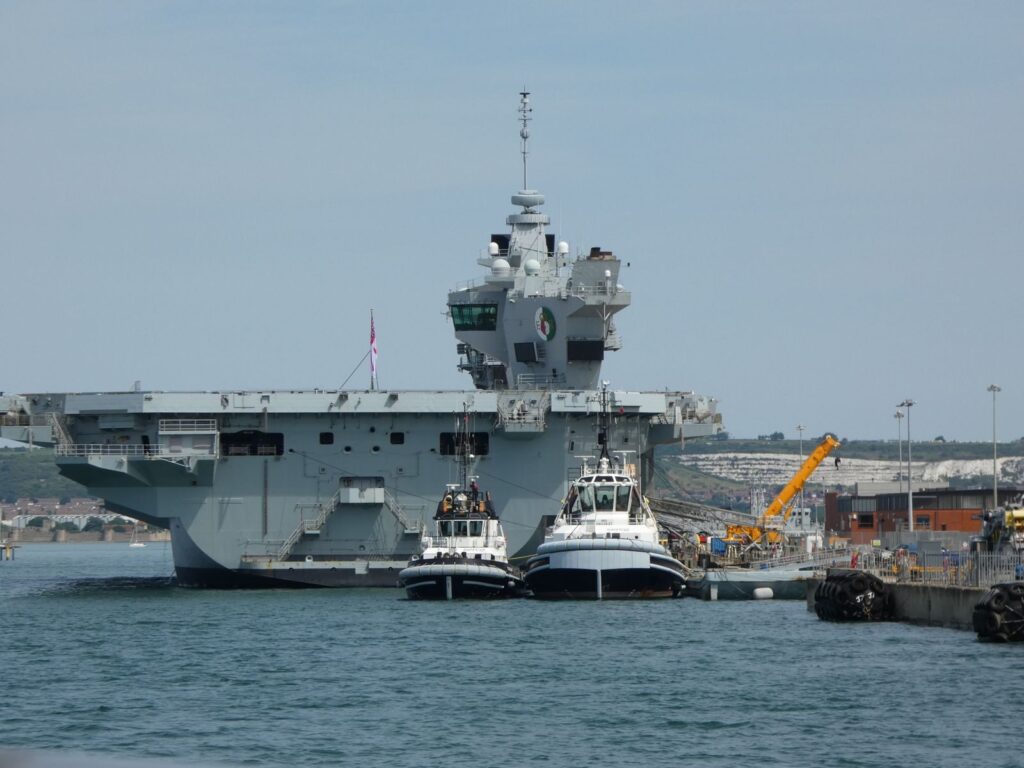 Impressive aircraft carrier docked at Portsmouth harbor with tugboats assisting.