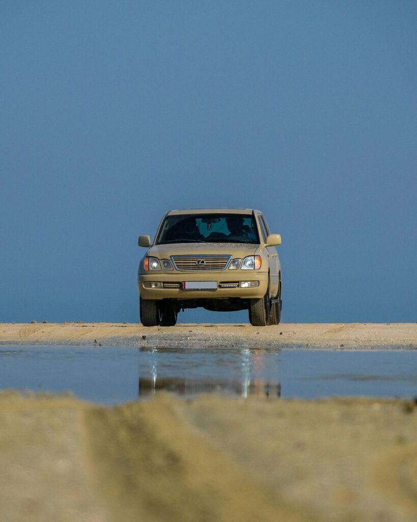 A Lexus SUV parked on a sandy terrain with a clear blue sky, reflecting in a puddle.
