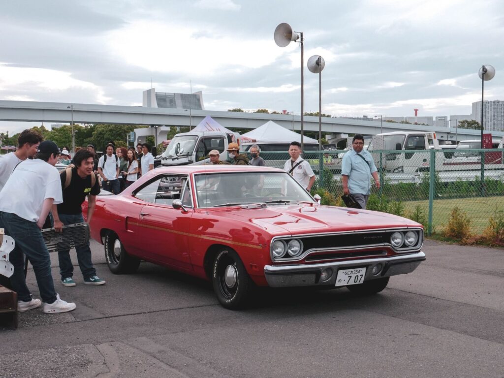 Red classic car showcased at an outdoor meetup with onlookers in summer.
