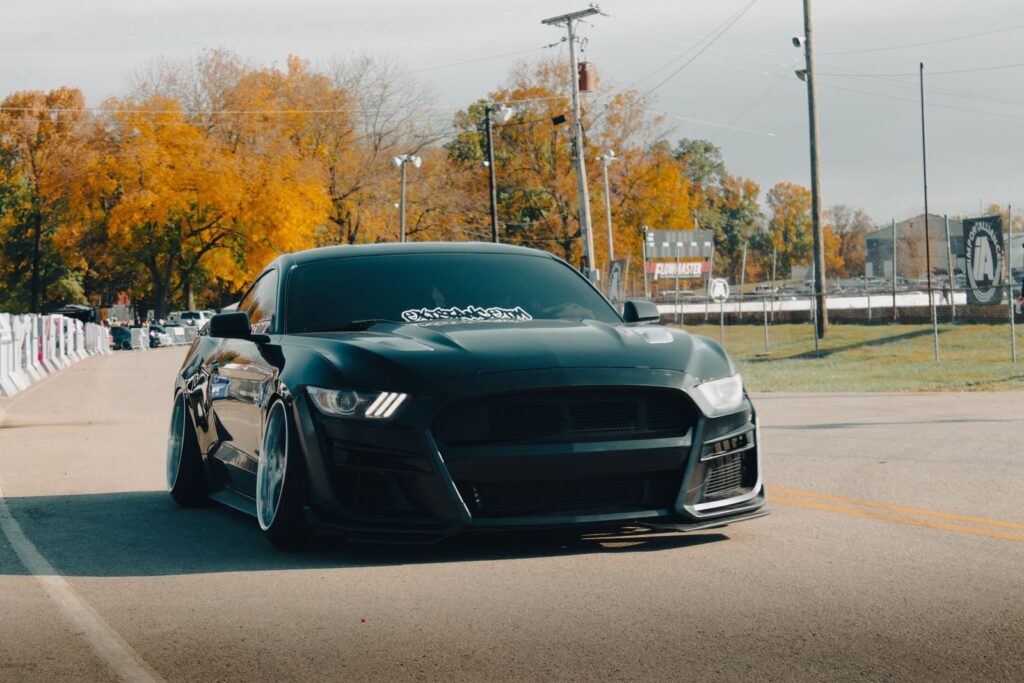 Stylish black Ford Mustang cruising at an outdoor car show with vibrant autumn leaves.