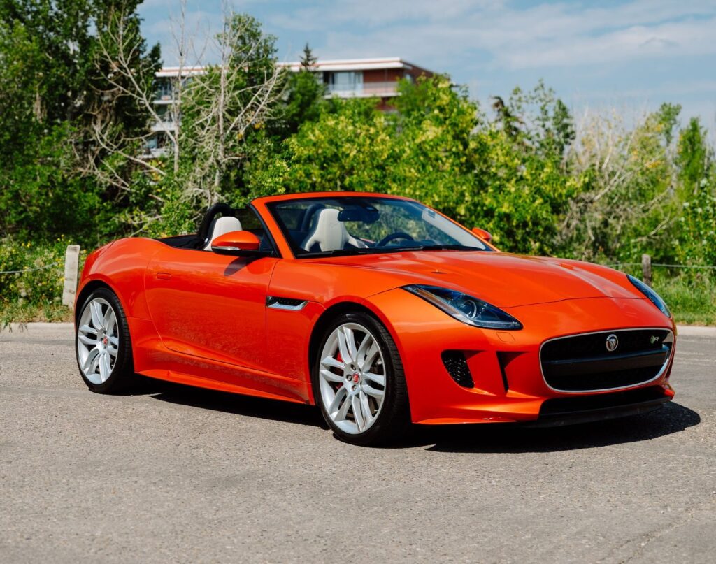 A bright orange Jaguar convertible parked outdoors under the sun, surrounded by lush greenery.