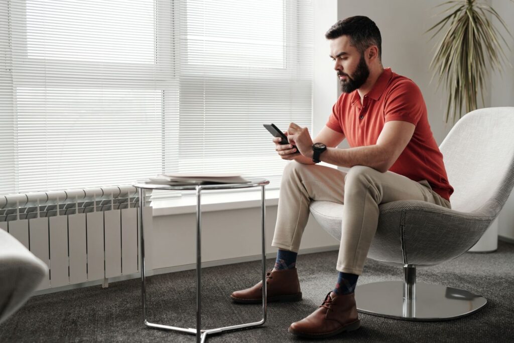 Bearded man in a red polo sitting on a modern chair using a smartphone, indoor setting.