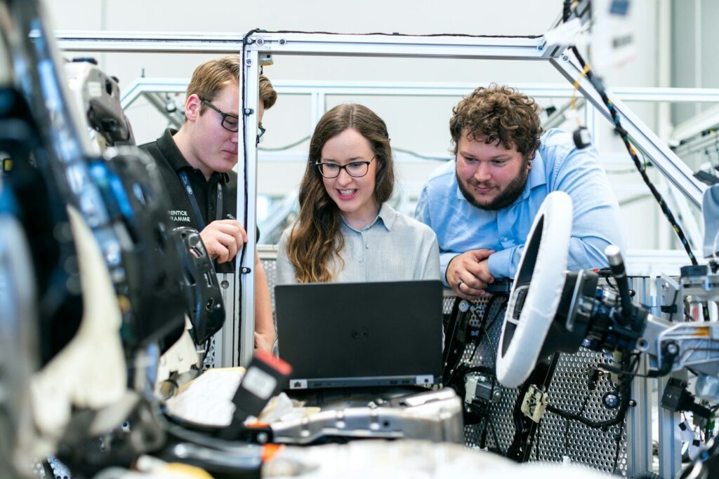 Three people looking at a laptop screen in an automotive assembly workspace